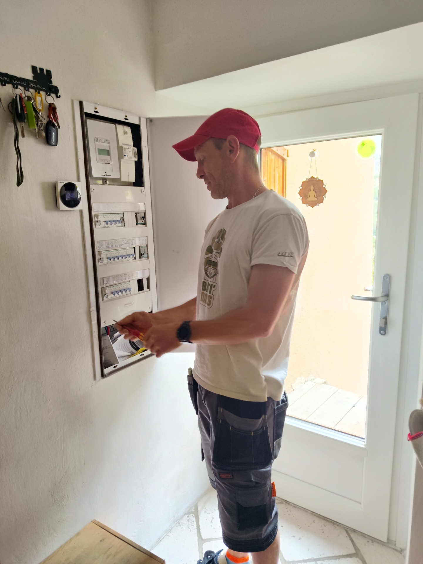 Homme en tenue de travail, casquette rouge, ajuste un tableau électrique près d’une porte vitrée.